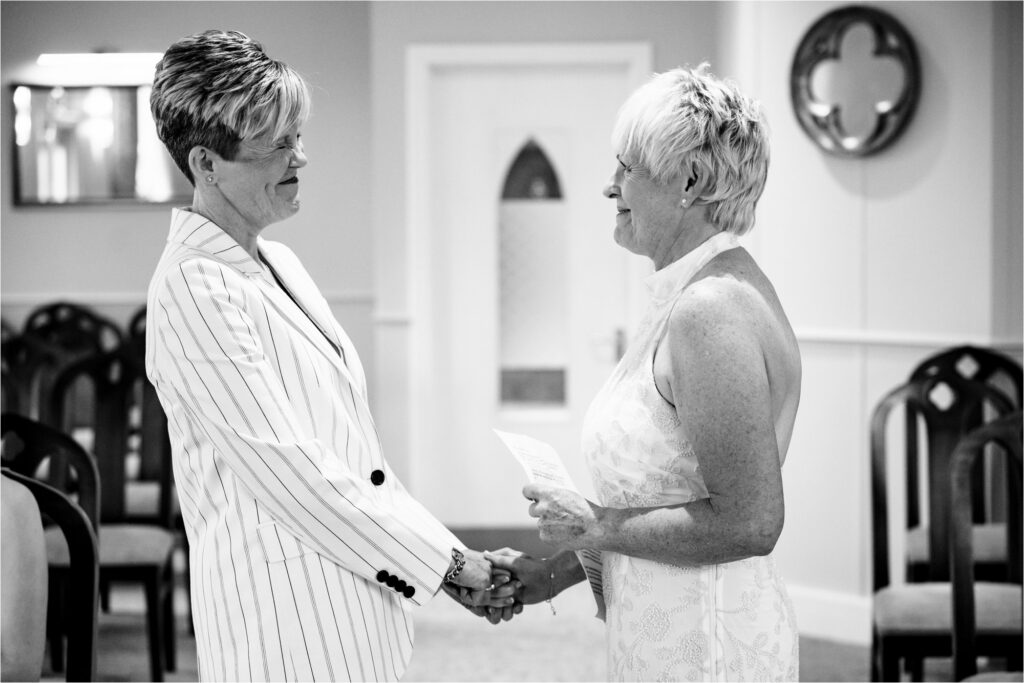 Bride in white pin stripe suit scrunches up her face in laughter during civil partnership vow exchange.