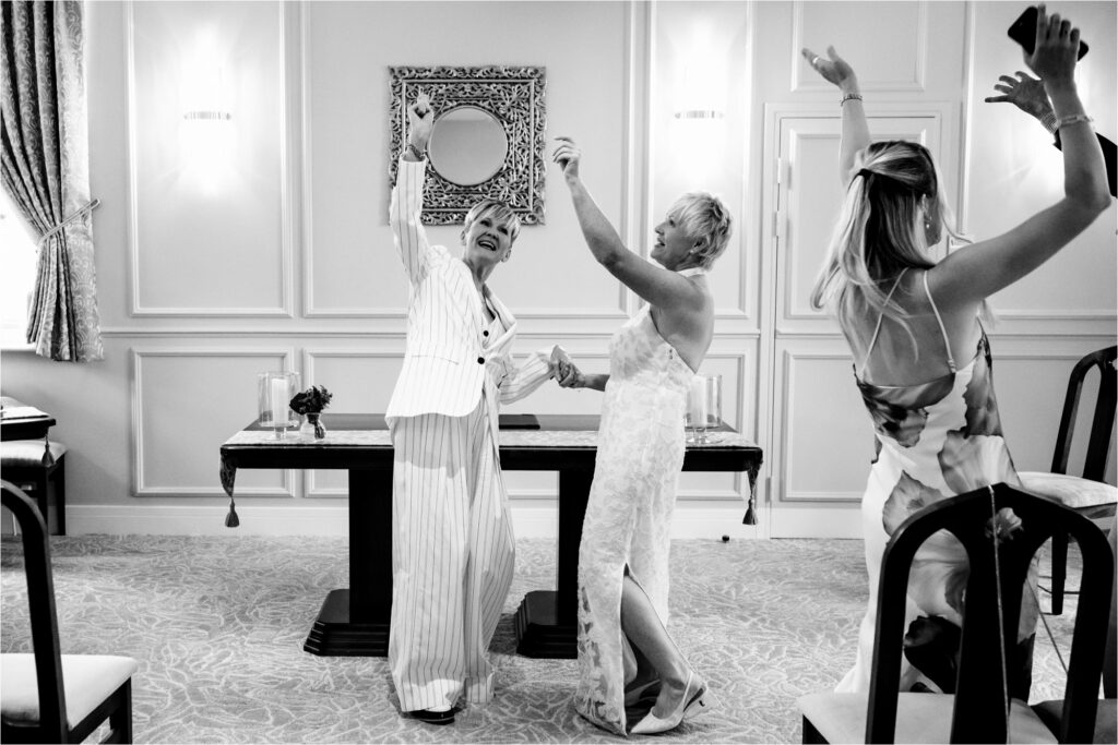Two brides and guests throw hands up in the air as they are pronounced married during registry office intimate wedding ceremony.