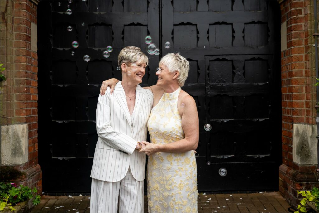 Two brides wearing a white suit and white dress stand in front of black doors smiling at each other as guests blow bubbles towards them.