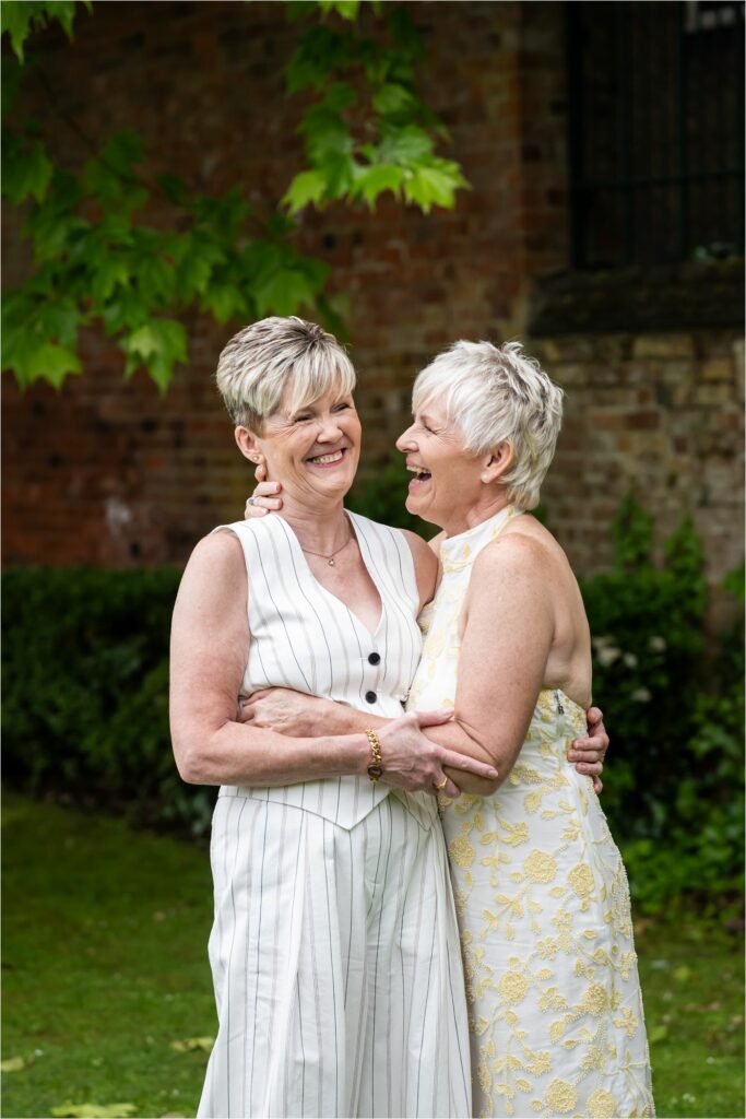 Two brides hug and laugh in front of summery green leafed tree in register office gardens.