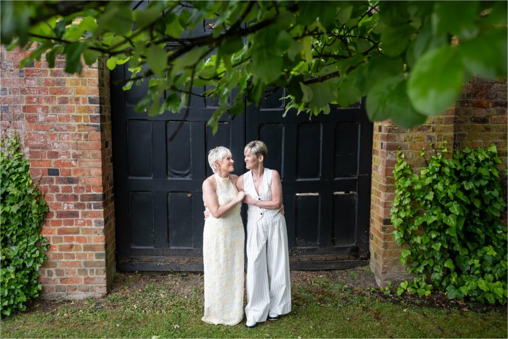 Two brides embrace under a green leafy tree in front of large black doors in gardens of St Albans Register Office.