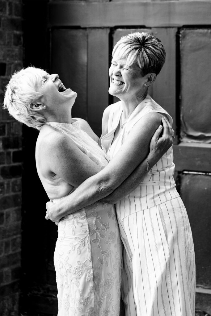 Two brides laugh hysterically with their eyes shut, holding each other in a tight embrace.