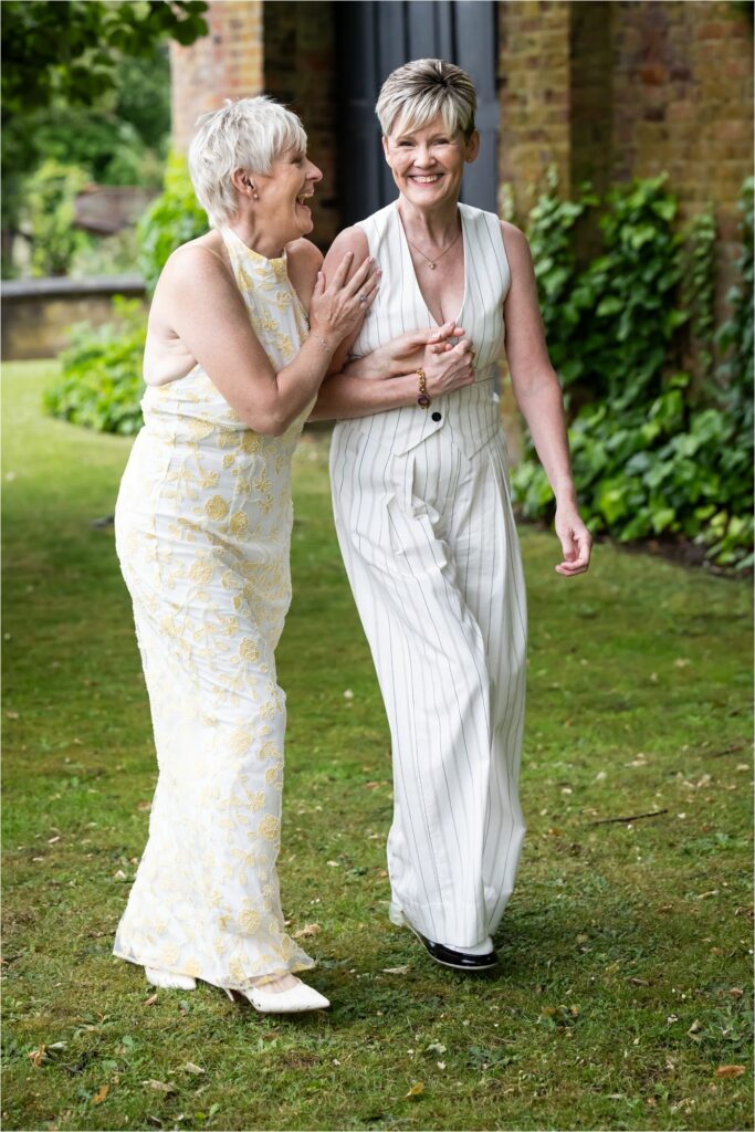 Two brides wearing a white suit and a white dress laugh and walk together through the St Albans Register Office gardens.