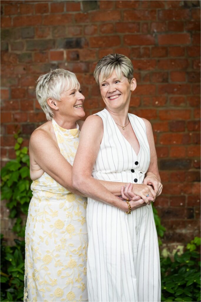 Bride in white patterned dress embraces her new wife in pinstripe trouser suit in front of red brick wall.