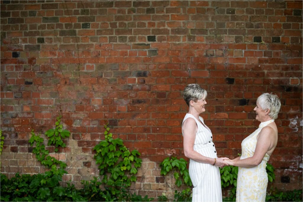 Laughing bride in white patterned dress faces her new wife in pinstripe trouser suit in front of red brick wall.