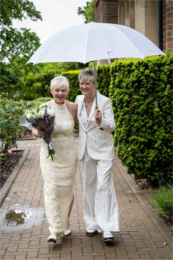 Two brides walk away from St Albans Register Office under a large white golfing umbrella holding a bouquet of flowers.