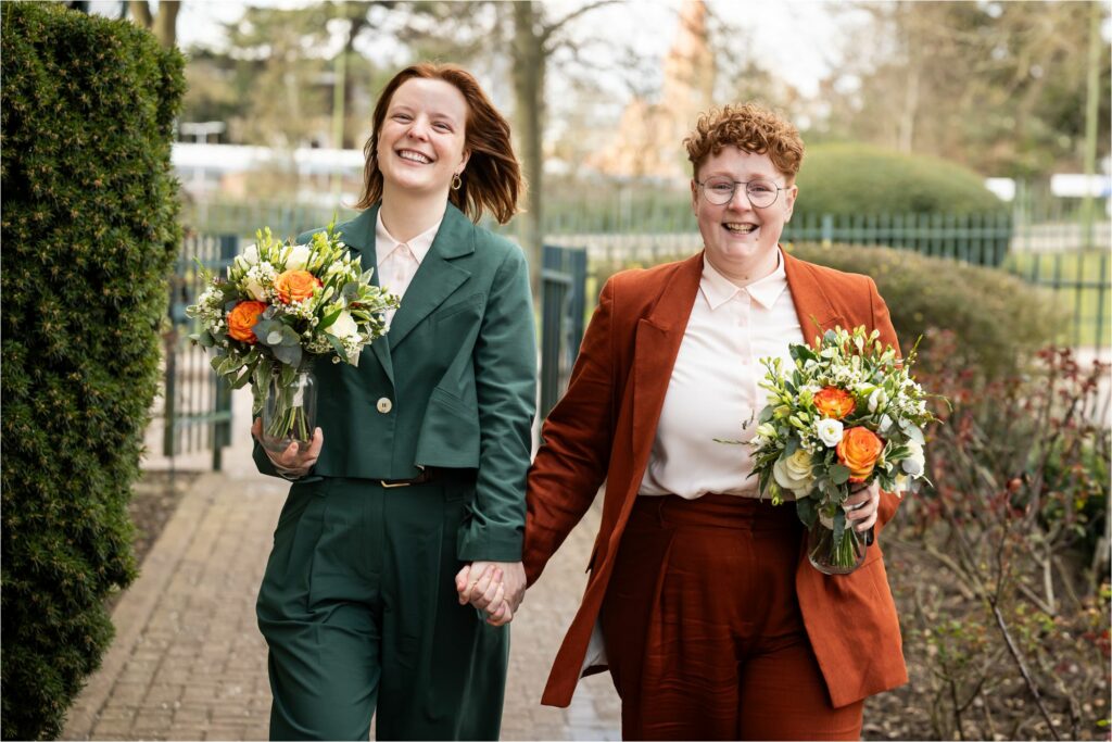 Two brides in suits, each holding a bouquet of flowers, walk hand-in-hand towards their wedding ceremony and smile at the camera.