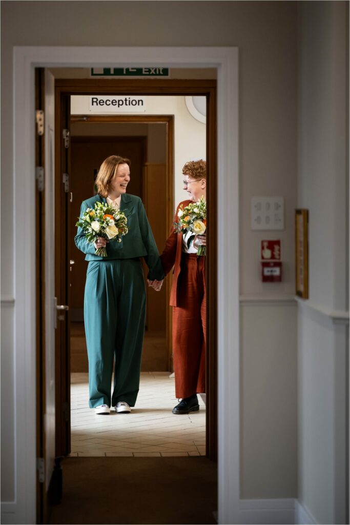 Two brides holding bouquets and hands, standing in the hallway waiting to be called into their wedding ceremony, grinning at each other.