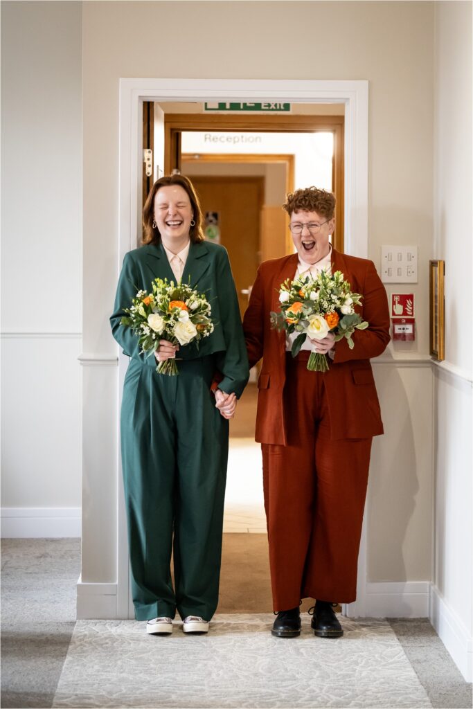 Two brides stand together holding hands at the reception room doorway, each holding a bouquet of flowers, dressed in stylish suits.
