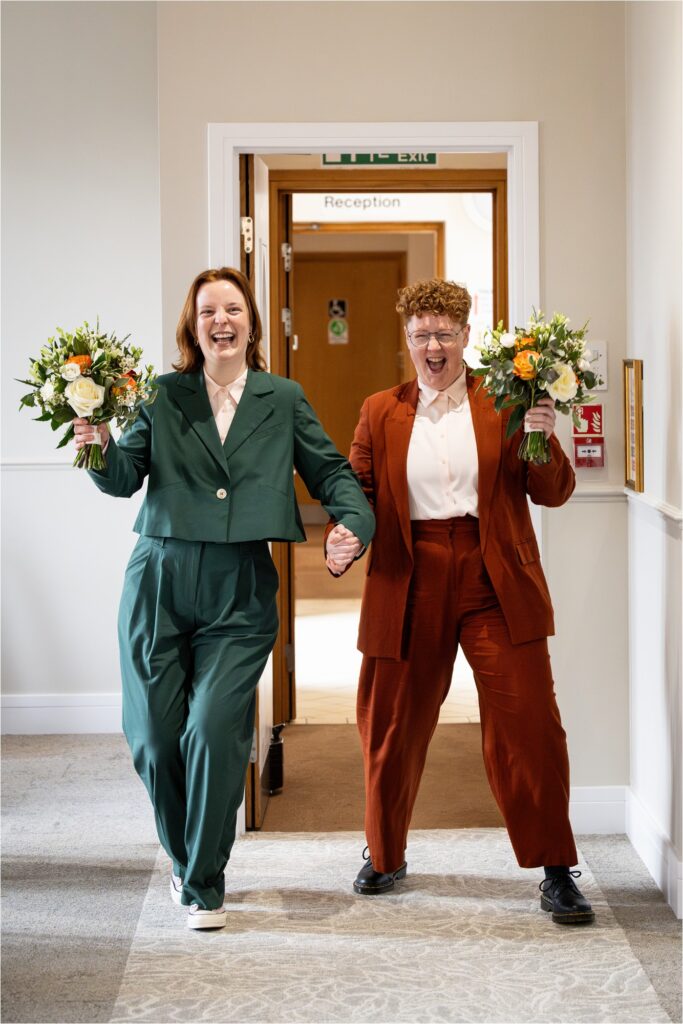 Two brides in stylish suits hold hands while carrying bouquets, dance confidently into their wedding ceremony.