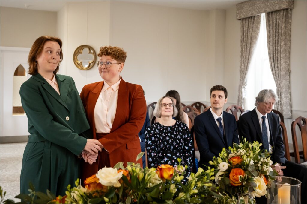 Two brides stand together, hands clasped, with a floral arrangement in the foreground of the St Albans Register Office.