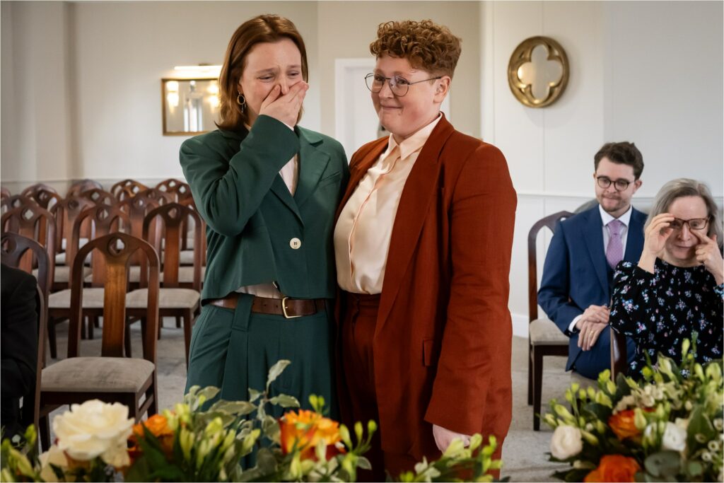Two brides in stylish suits stand together, in an intimate ceremony, with floral decorations in front.