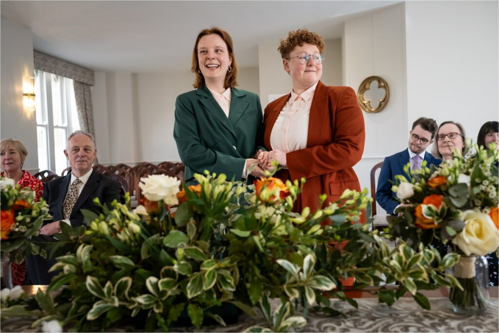 Two brides engage in a heartfelt moment holding hands at a floral-adorned ceremony, with seated guests in the background.