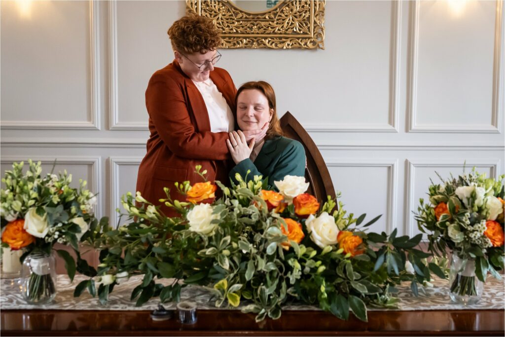 Two brides share a heartfelt hug at a wedding table filled with colourful floral arrangements.