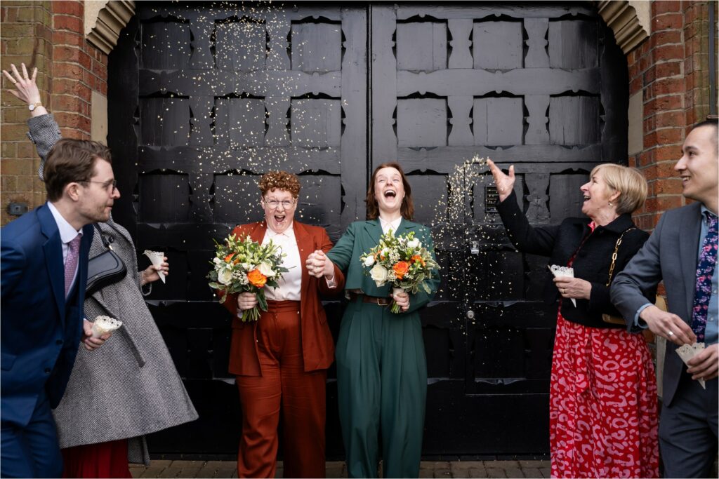 Family throw confetti over two brides leaving the St Albans Registry Office in front of black doors.