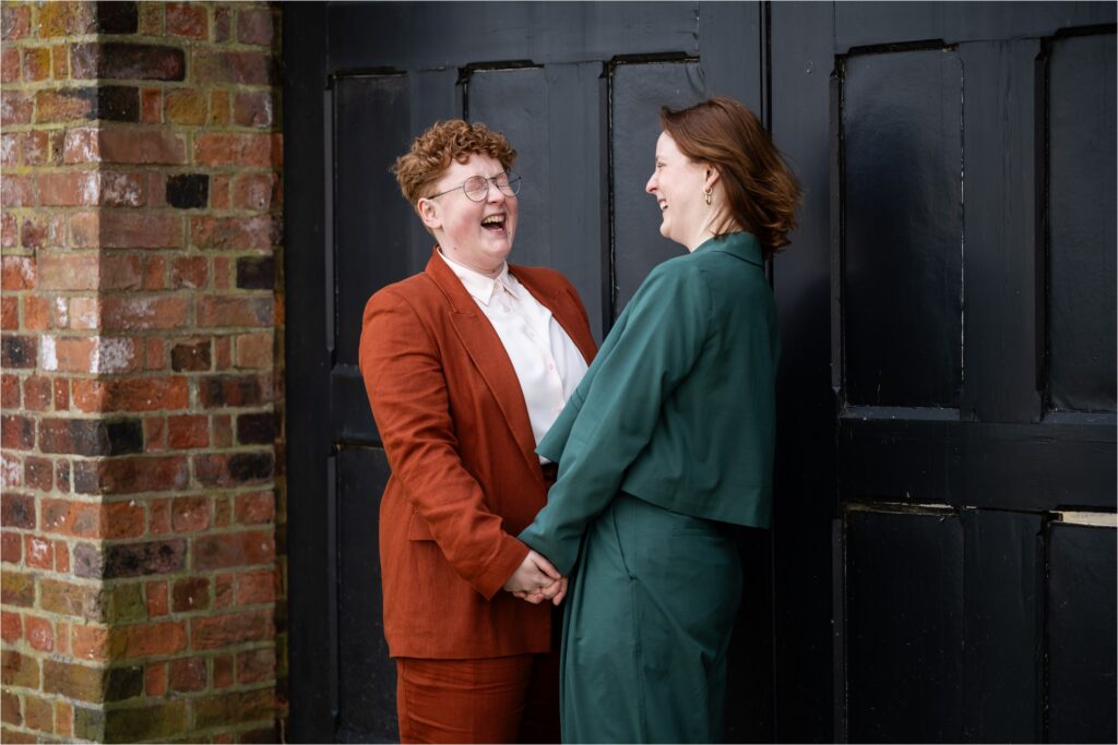 Two brides in colourful suits laugh in front of black doors in St Albans Register Office gardens.