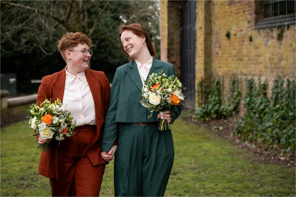 Two brides hold hands and carry bouquets and smile at each other walking through St Albans Register Office gardens.