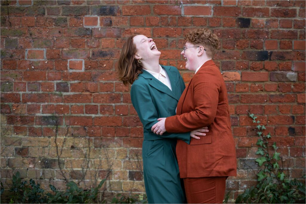 Two laughing brides wearing suits embrace in front of the St Albans Register Office red walls.