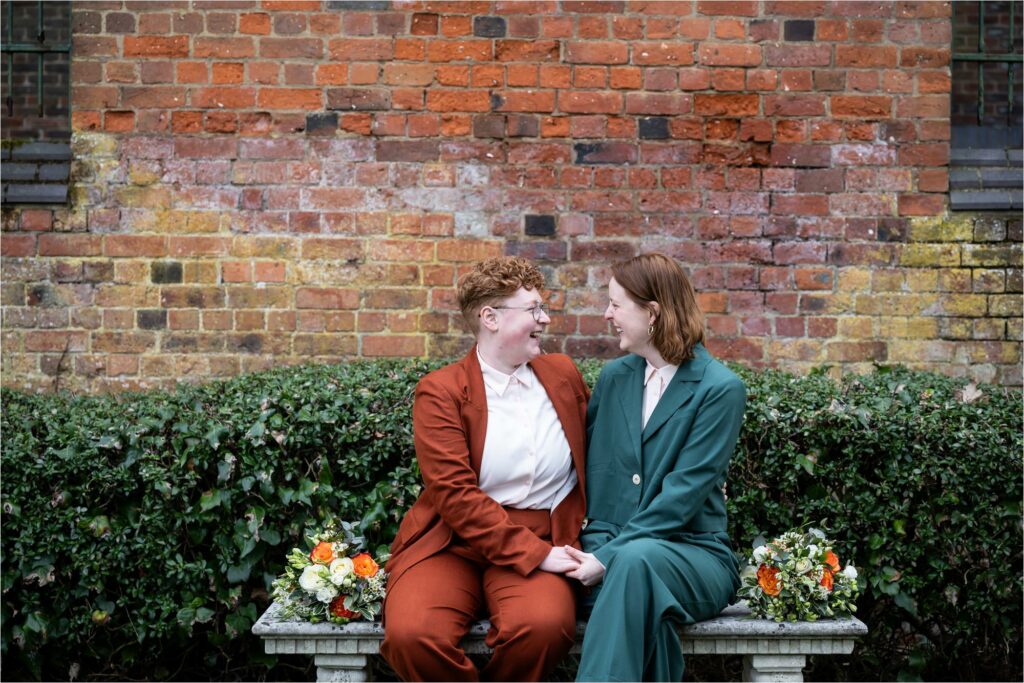 Two brides sit on a stone bench holding hands and smile at each other in St Albans Register Office.