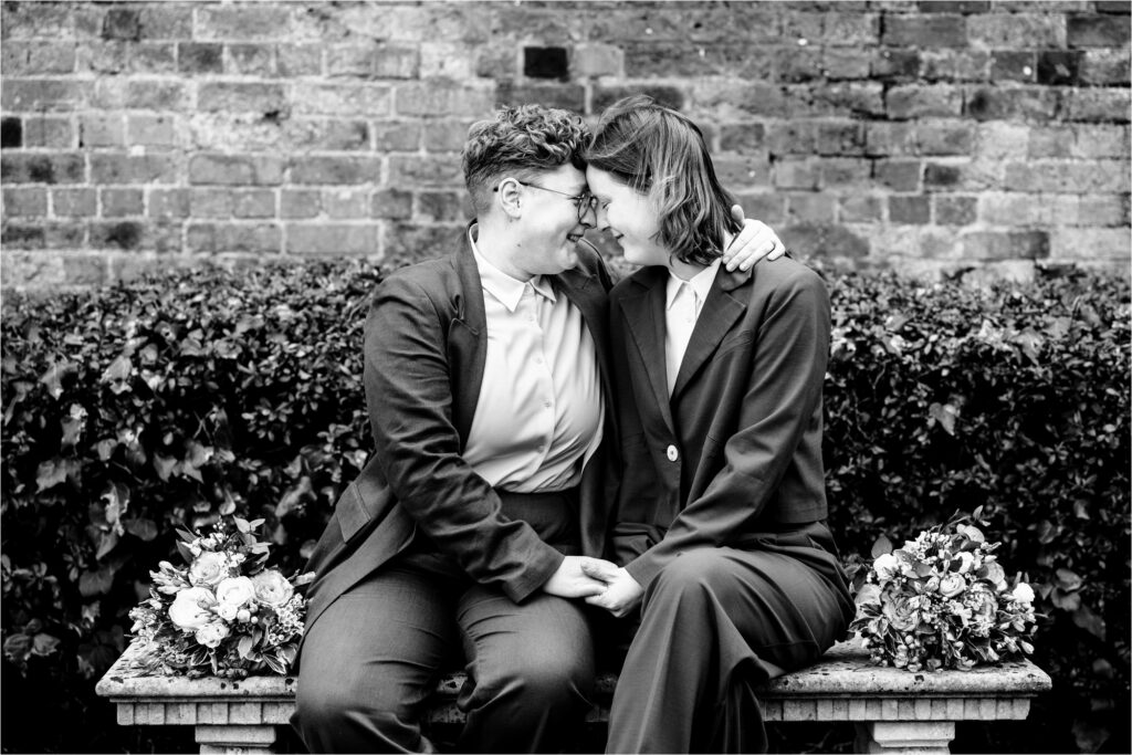 Two brides wearing suits touch foreheads in peaceful moment sitting on a bench in a garden.